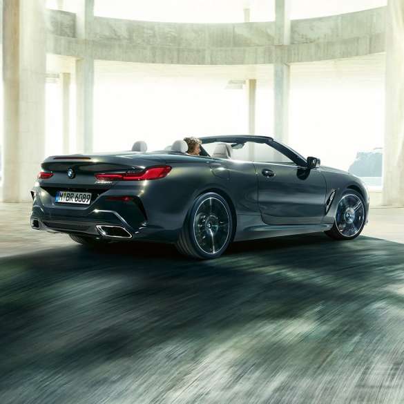 BMW M850i xDrive Cabrio, tříčtvrteční pohled zezadu. BMW M850i xDrive Cabrio v šedé metalíze Dravit Grey se staženou střechou, tříčtvrteční pohled zezadu na vůz projíždějící betonovou budovou.