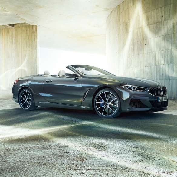 BMW M850i xDrive Cabrio, tříčtvrteční pohled z boku. BMW M850i xDrive Cabrio v šedé metalíze Dravit Grey se staženou střechou, tříčtvrteční pohled z boku, stojící před betonovou budovou.
