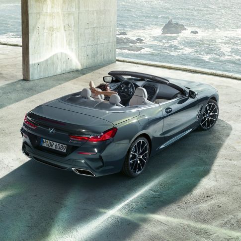 BMW M850i xDrive Cabrio, pohled z ptačí perspektivy. BMW M850i xDrive Cabrio v šedé metalíze Dravit Grey se staženou střechou, pohled z ptačí perspektivy na vůz zaparkovaný v betonové budově před oceánem.