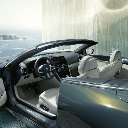 BMW M850i xDrive Cabrio, interiér. BMW M850i xDrive Cabrio v šedé metalíze Dravit Grey se staženou střechou, interiér s M koženým volantem a sedadly čalouněnými bílou kůží BMW Individual Merino Ivory White.