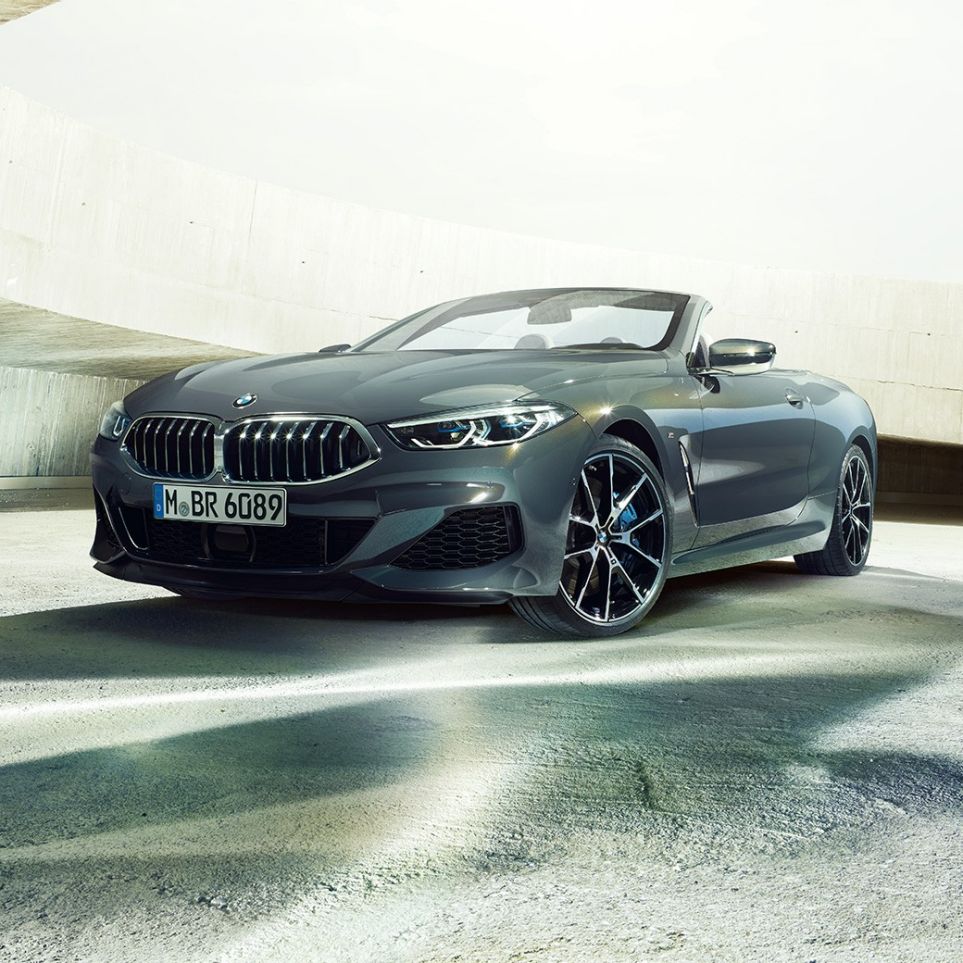 BMW M850i xDrive Cabrio, tříčtvrteční pohled zepředu. BMW M850i xDrive Cabrio v šedé metalíze Dravit Grey se staženou střechou, tříčtvrteční pohled zepředu na vůz stojící v betonové budově.