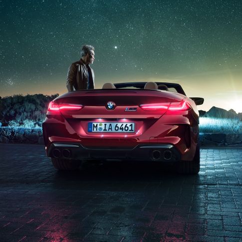 BMW M8 Competition Cabrio, pohled zezadu. BMW M8 Copmpetition Cabrio v červené metalíze Motegi Red se staženou střechou, pohled zezadu s mužem stojícím pod hvězdnou oblohou.