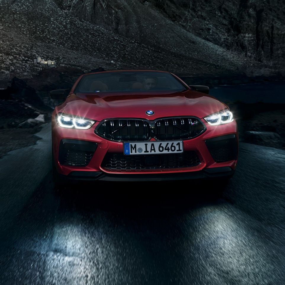 BMW M8 Competition Cabrio, pohled zepředu. BMW M8 Competition Cabrio v červené metalíze Motegi Red se zářícími předními světlomety, jízdní pohled v noci zepředu.