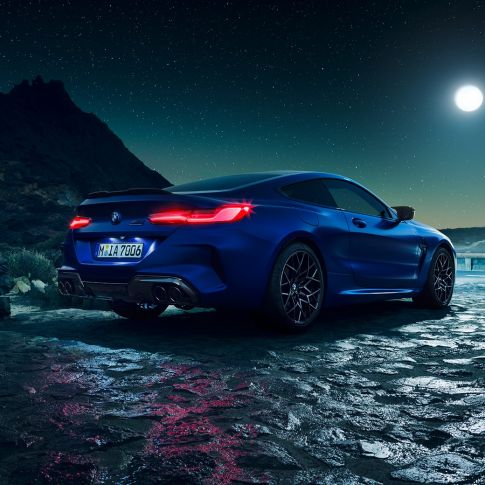 BMW M8 Competition Coupé, tříčtvrteční pohled zezadu. BMW M8 Competition Coupé v metalické barvě Marina Bay Blue s rozsvícenými LED zadními světly, tříčtvrteční pohled zezadu na pozadí úplňku.