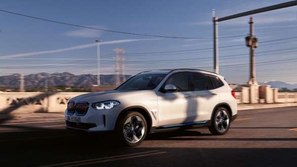 Adaptivní M podvozek BMW ZiX3 G08 jedoucí na ulici Adaptivní M podvozek BMW iX3 G08 2020 v metalické barvě Mineral White (bílá) z tříčtvrtečního pohledu jedoucí na ulici
