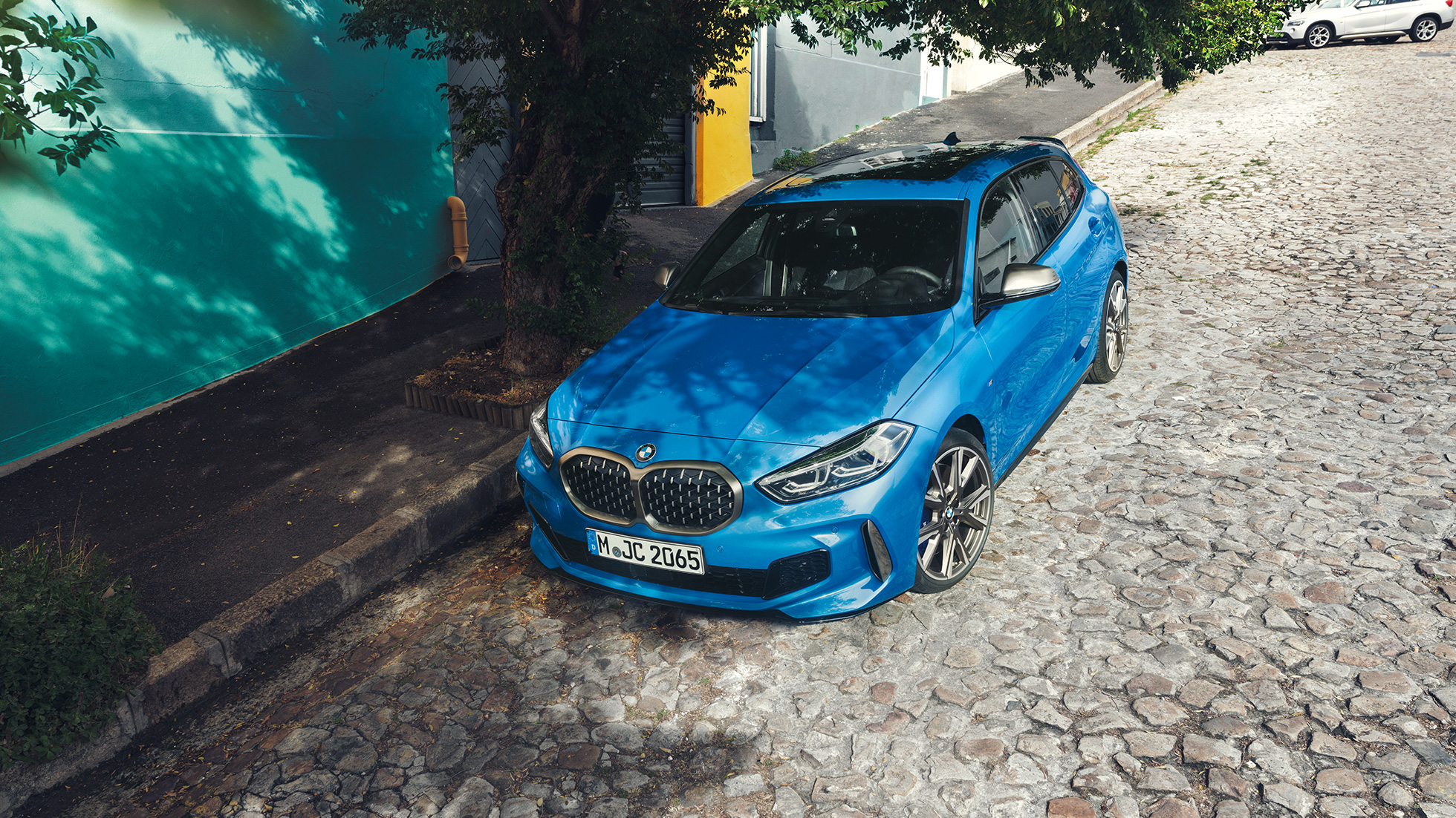 BMW řady 1 na krajnici. Tříčtvrteční pohled z ptačí perspektivy na BMW řady 1 v modré metalické barvě Misano Blue, zaparkované na krajnici.