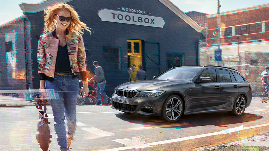 BMW řady 3 Touring (G21) tříčtvrteční záběr zepředu se ženou před dřevařským obchodem. Žena přechází ulici před BMW řady 3 Touring (G21) před dřevařským obchodem.
