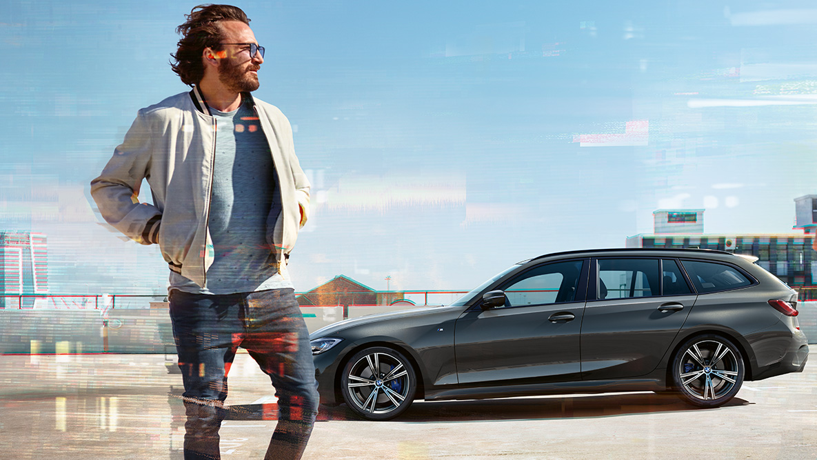BMW řady 3 Touring (G21), tříčtvrteční pohled zepředu BMW řady 3 Touring (G21) v šedé barvě Dravit Grey, jízdní tříčtvrteční záběr zepředu na mostě ve venkovském prostředí