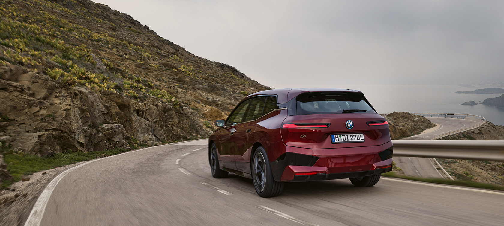 BMW iX i20 2021 elektrické SUV BMW iX xDrive50 při jízdě po pobřeží BMW iX i20 2021 elektrické SUV BMW iX xDrive50 Aventurin Red (červená) pohled zezadu při jízdě po pobřeží