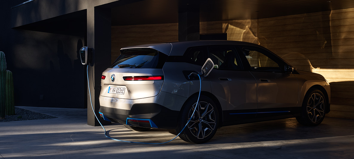 BMW iX tříčtvrteční pohled zezadu při nabíjení nabíječkou BMW Wallbox BMW iX Cashmere Silver i20 tříčtvrteční pohled zezadu při nabíjení nabíječkou BMW Wallbox