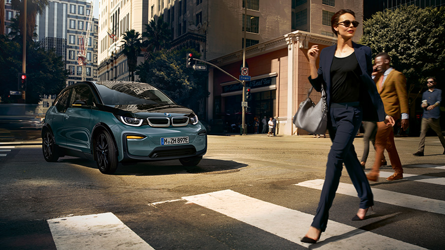 BMW i3 I01 tříčtvrteční pohled zepředu se ženou přecházející přes přechod BMW i3 I01 v modré barvě Blue Ridge Mountain s matně šedými kontrastními prvky Frozen Grey tříčtvrteční pohled zepředu se ženou přecházející přes přechod