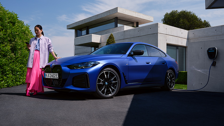 BMW i4 M50 G26 2021 metalick&yacute; lak Individual Frozen Portimao Blue (modr&aacute;) tř&iacute;čtvrtečn&iacute; pohled zepředu se ženou stoj&iacute;c&iacute; před domem