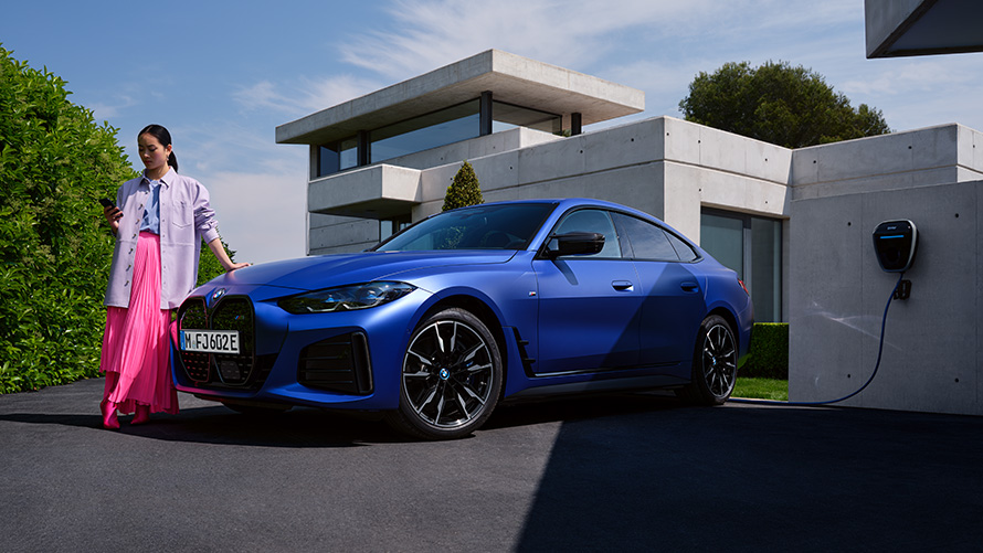 Tříčtvrteční pohled z boku BMW i4 M50 G26 2021 se ženou před domem BMW i4 M50 G26 2021 metalický lak Individual Frozen Portimao Blue (modrá) tříčtvrteční pohled z boku se ženou před domem