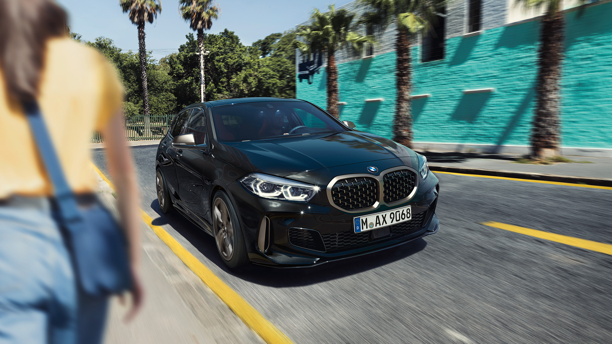 BMW M135i xDrive, tříčtvrteční pohled zepředu. BMW M135i xDrive v černé metalíze Black Sapphire, tříčtvrteční pohled zepředu s ženou v popředí.