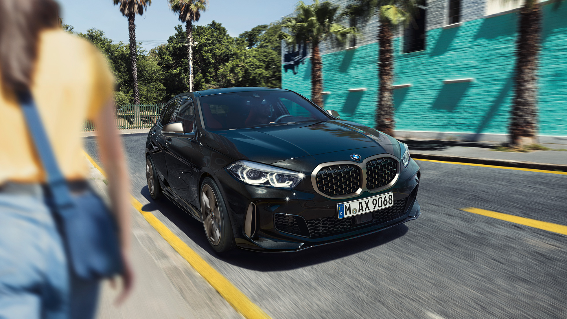 BMW M135i xDrive, tříčtvrteční pohled zepředu. BMW M135i xDrive v černé metalíze Black Sapphire, tříčtvrteční pohled zepředu s ženou v popředí.