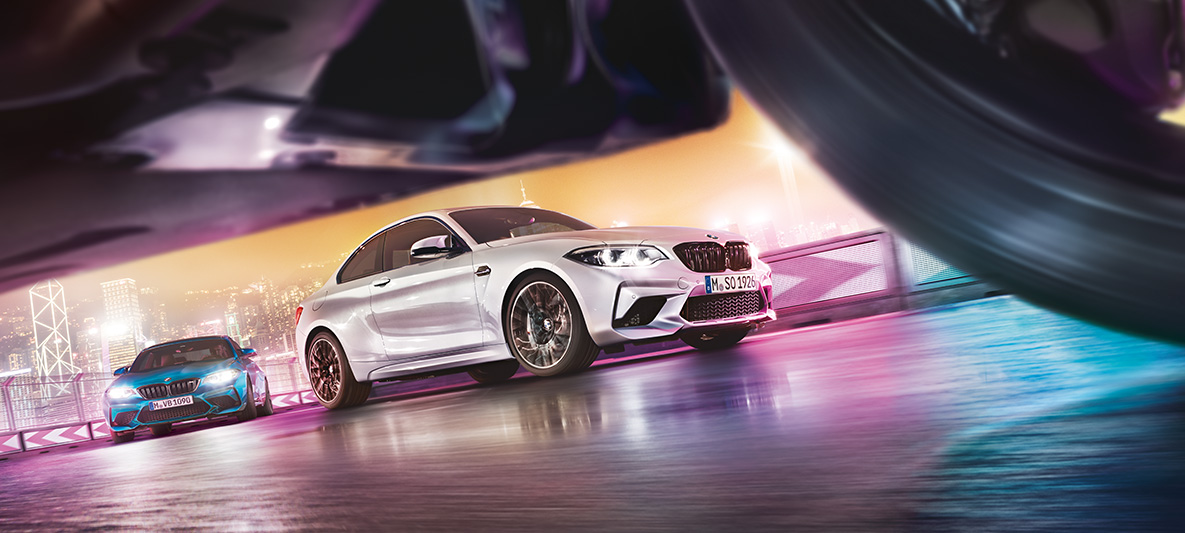 BMW M2 Competition na reflexním asfaltu. Dvě BMW M2 Competition driftující po závodní dráze s panoramatem nočního města v pozadí z úrovně ulice.