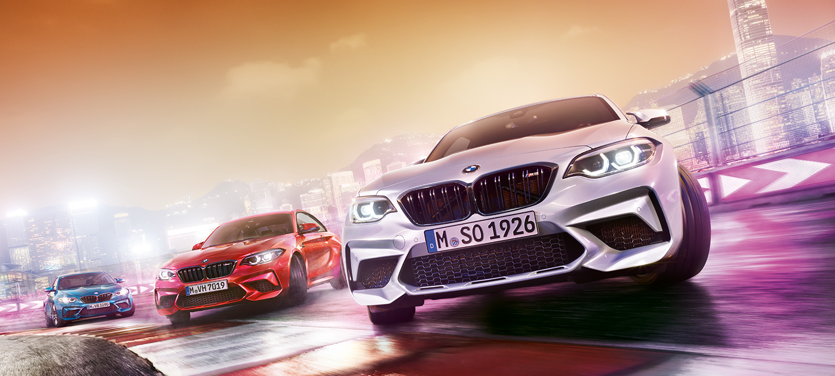 BMW M2 Competition v akci Tři BMW M2 Competition driftující po závodní dráze s panoramatem nočního města v pozadí.