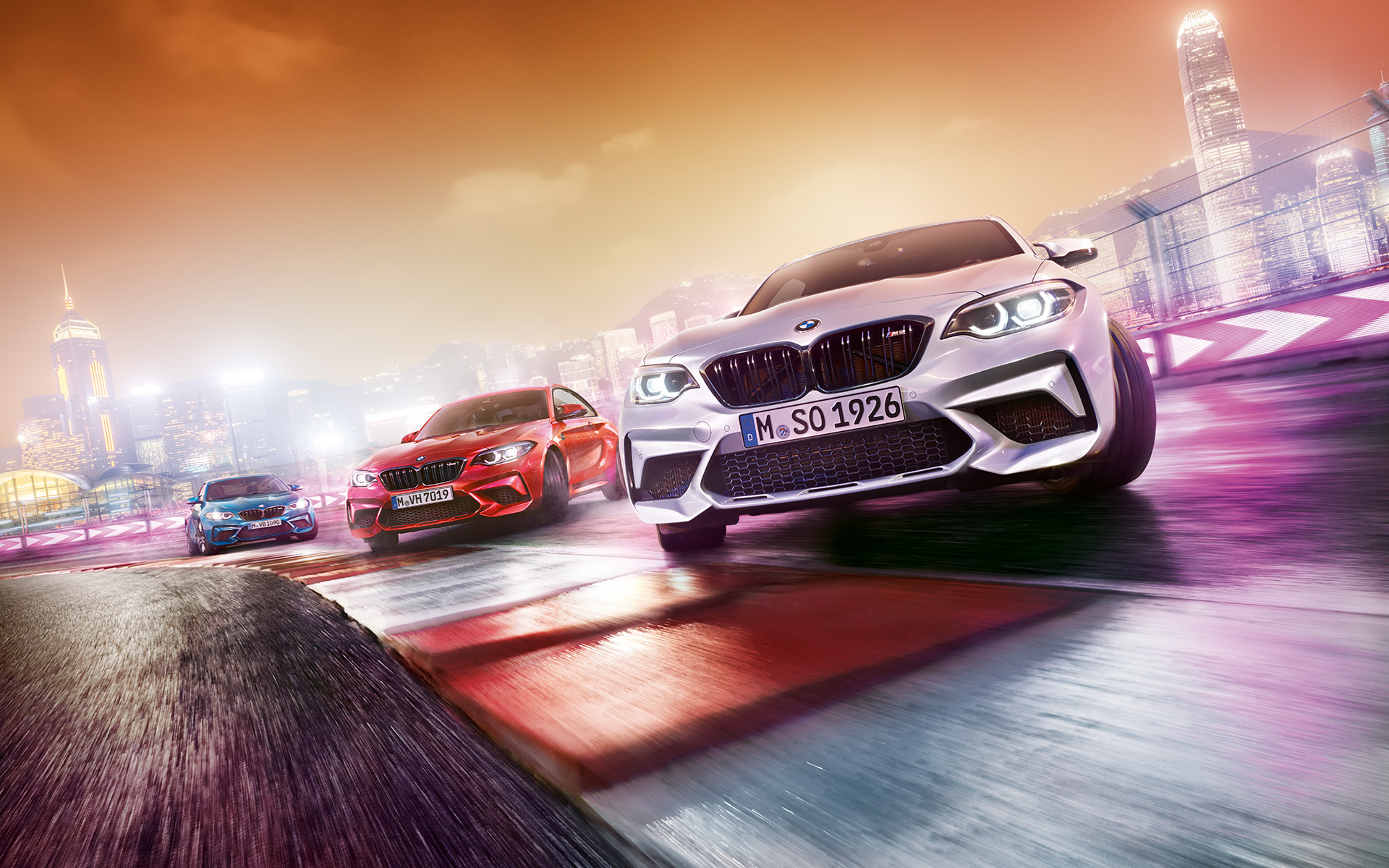 BMW M2 Competition v akci Tři BMW M2 Competition driftující na závodní dráze s panoramatem nočního města v pozadí.