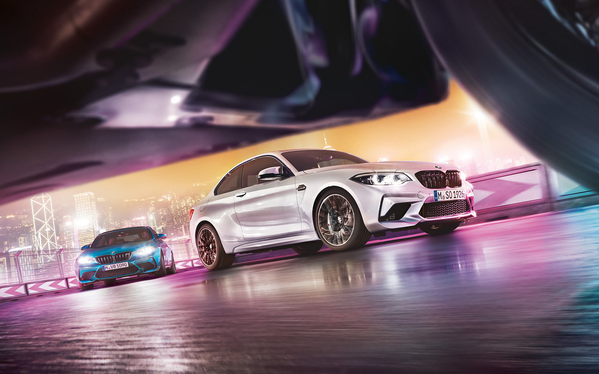 BMW M2 Competition na reflexním asfaltu Dvě BMW M2 Competition na závodní dráze s panoramatem nočního města v pozadí.