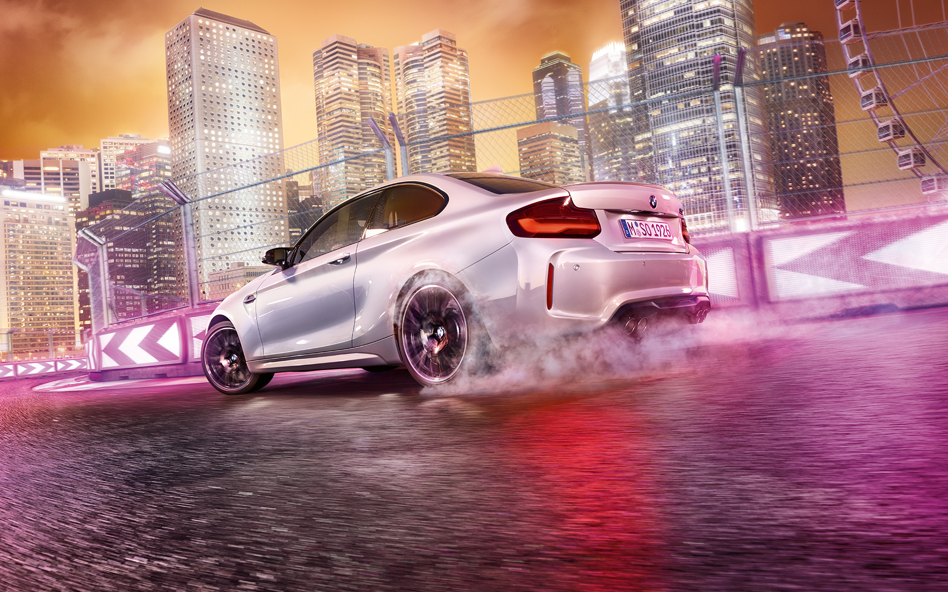 BMW M2 Competition zrychluje Bílé BMW M2 Competition driftuje po závodní dráze s panoramatem nočního města v pozadí.