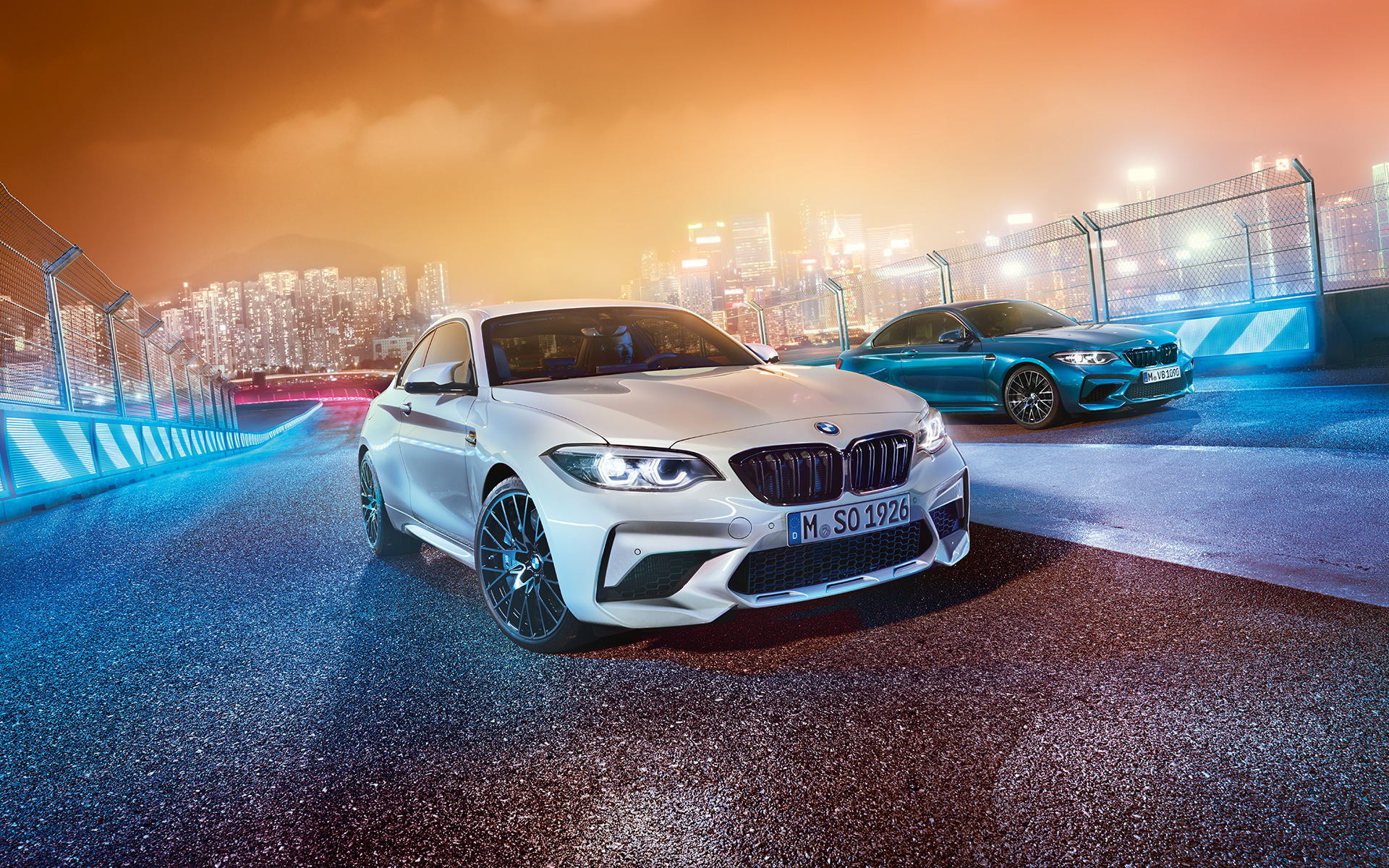 Dvě BMW M2 Competition na závodní trati Bílé a modré BMW M2 Competition stojí šikmo s panoramatem nočního města v pozadí.