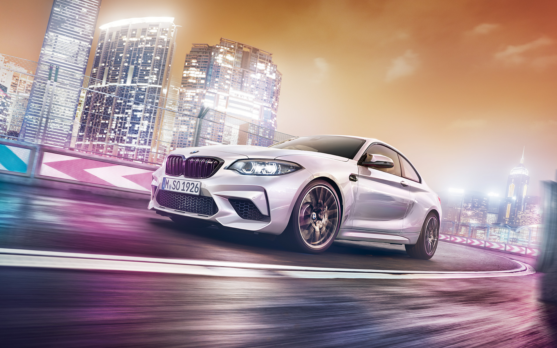 BMW M2 Competition s panoramatem města Bílé BMW M2 Competition jezdí po závodní trati s panoramatem nočního města v pozadí.