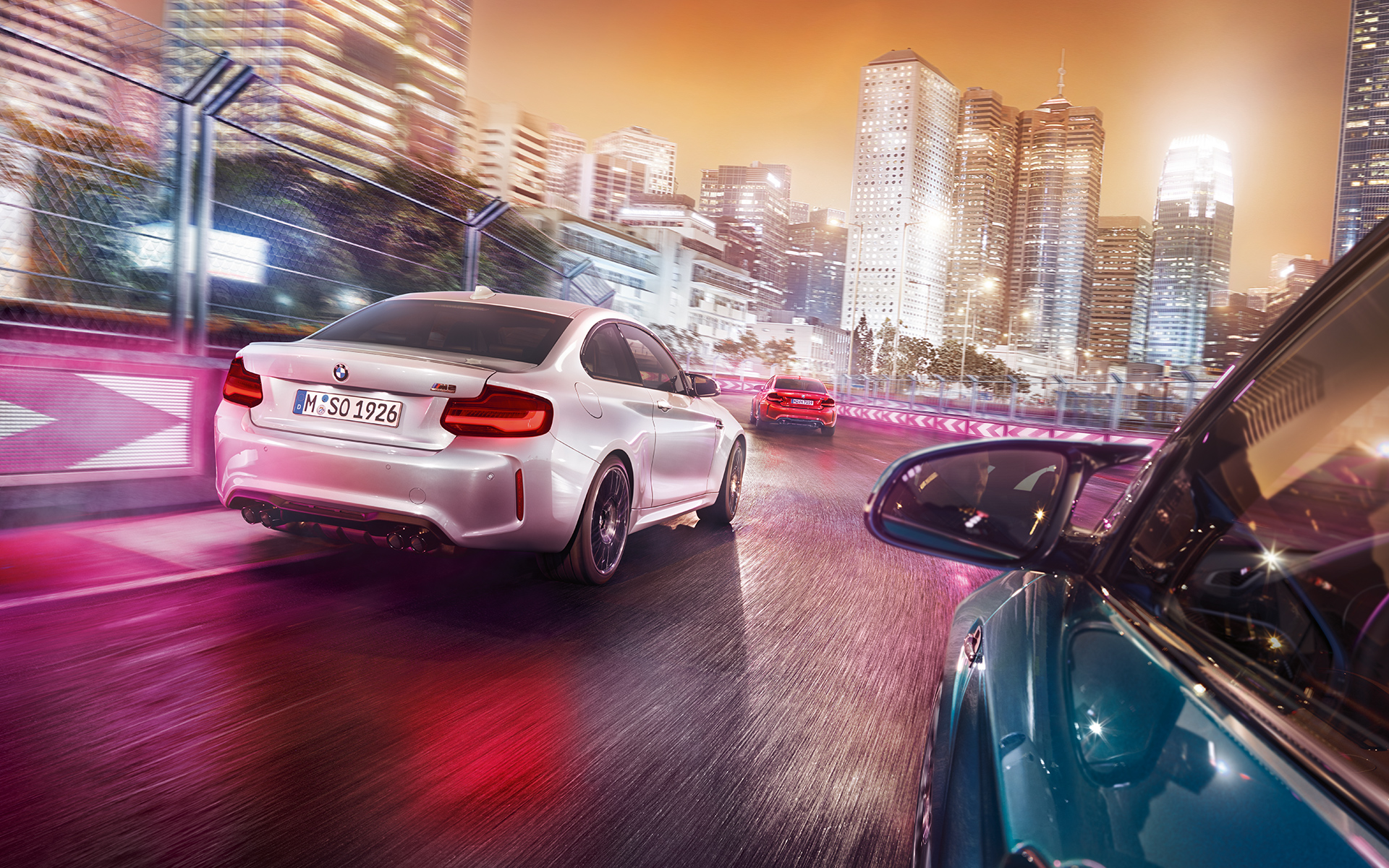 BMW M2 Competition pohled zezadu Tři BMW M2 Competition stojící šikmo zezadu na závodní trati s panoramatem nočního města v pozadí.