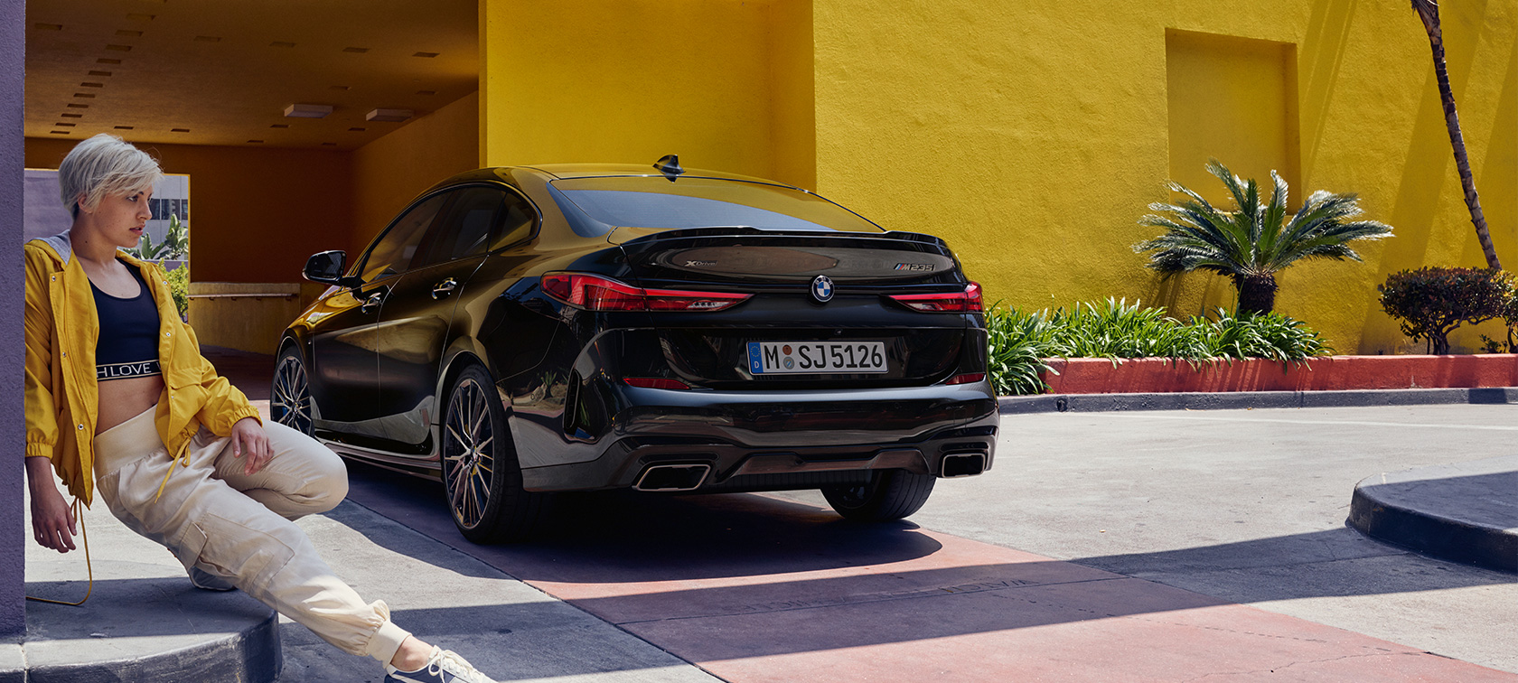 BMW M235i xDrive Gran Coupé ze tříčtvrtečního pohledu BMW M235i xDrive Gran Coupé F44 v metalické barvě Black Sapphire ze tříčtvrtečního pohledu zezadu se ženou vpředu