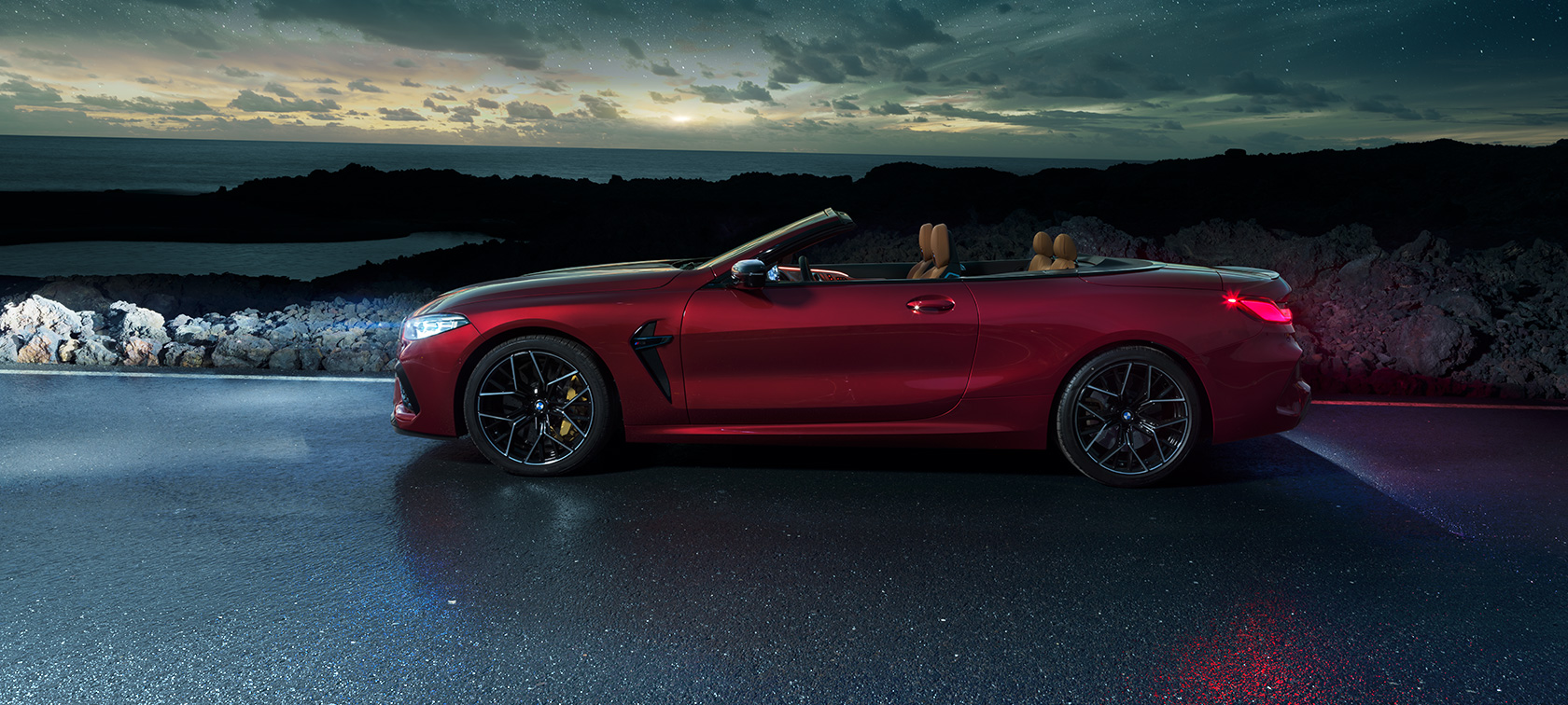 BMW M8 Competition Cabrio, boční pohled. BMW M8 Competition Cabrio v červené metalíze Motegi Red se staženou střechou, pohled z boku na vůz stojící na pobřežní silnici pod hvězdnou oblohou.