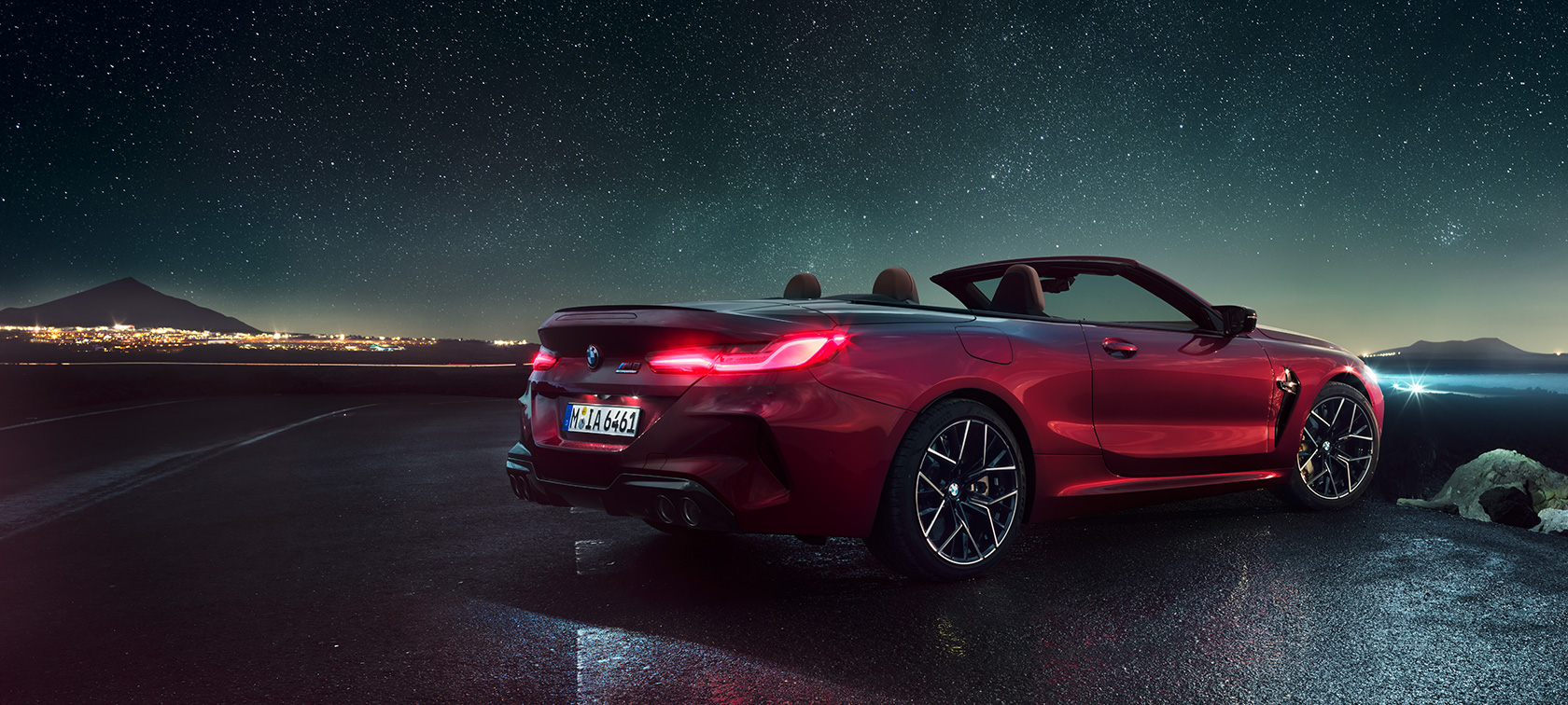 BMW M8 Competition Cabrio, tříčtvrteční pohled zezadu. BMW M8 Competition Cabrio v červené metalíze Motegi Red se staženou střechou, tříčtvrteční pohled zezadu na vůz stojící pod hvězdnou oblohou.
