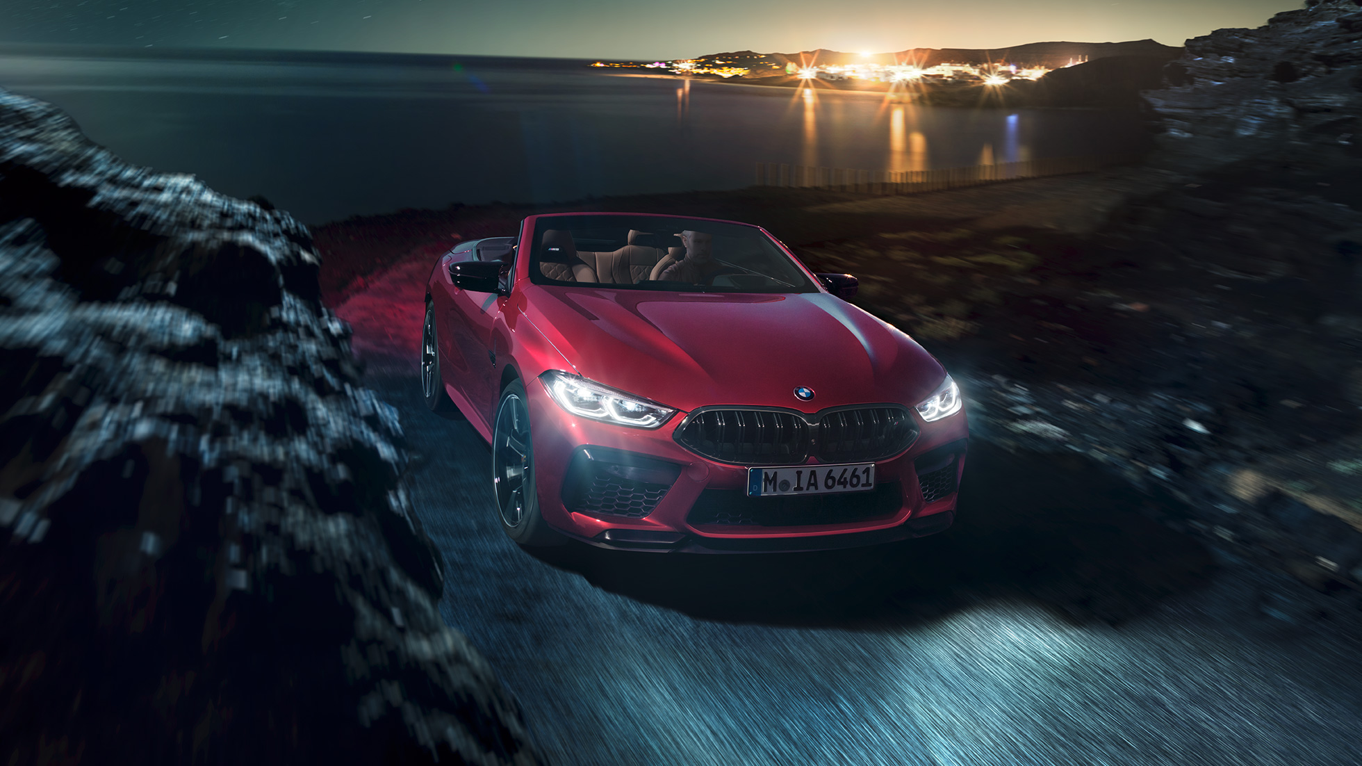 BMW M8 Competition Cabrio, tříčtvrteční pohled zepředu. BMW M8 Competition Cabrio v červené metalíze Motegi Red se staženou střechou, tříčtvrteční noční pohled zepředu na vozidlo jedoucí po pobřeží.