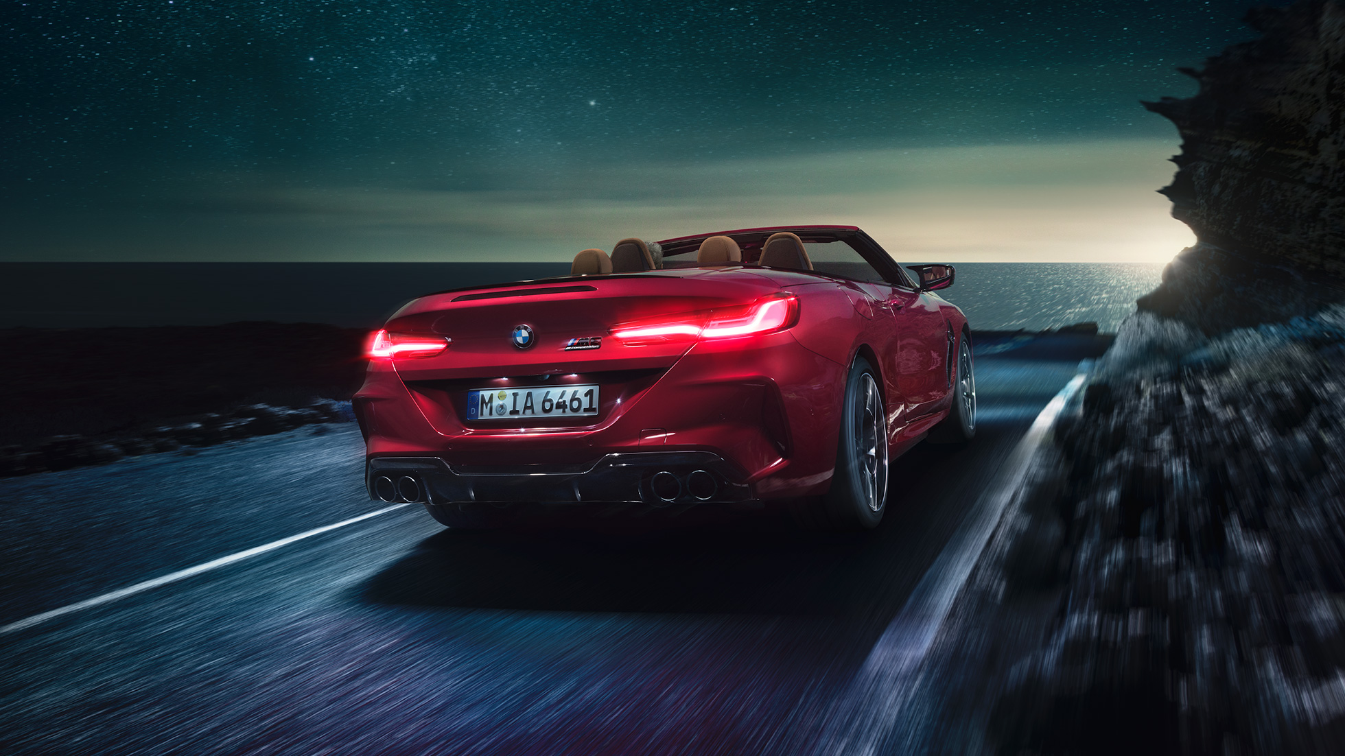 BMW M8 Competition Cabrio, tříčtvrteční pohled zezadu. BMW M8 Competition Cabrio v červené metalíze Motegi Red se staženou střechou, tříčtvrteční noční pohled zezadu na vozidlo jedoucí po pobřeží.