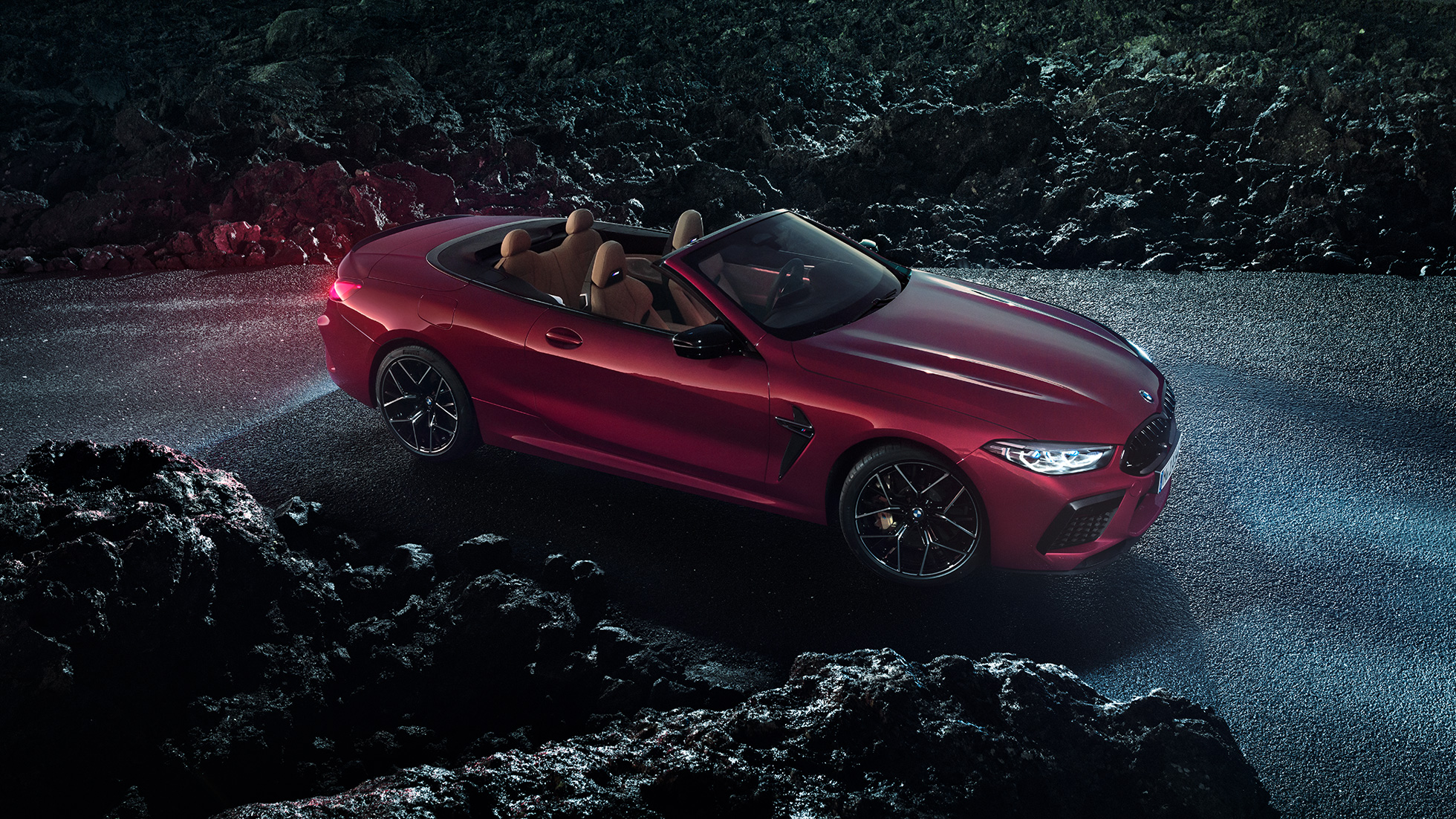 BMW M8 Competition Cabrio, tříčtvrteční boční pohled. BMW M8 Competition Cabrio v červené metalíze Motegi Red se staženou střechou, tříčtvrteční noční pohled z boku na vozidlo stojící na asfaltu.