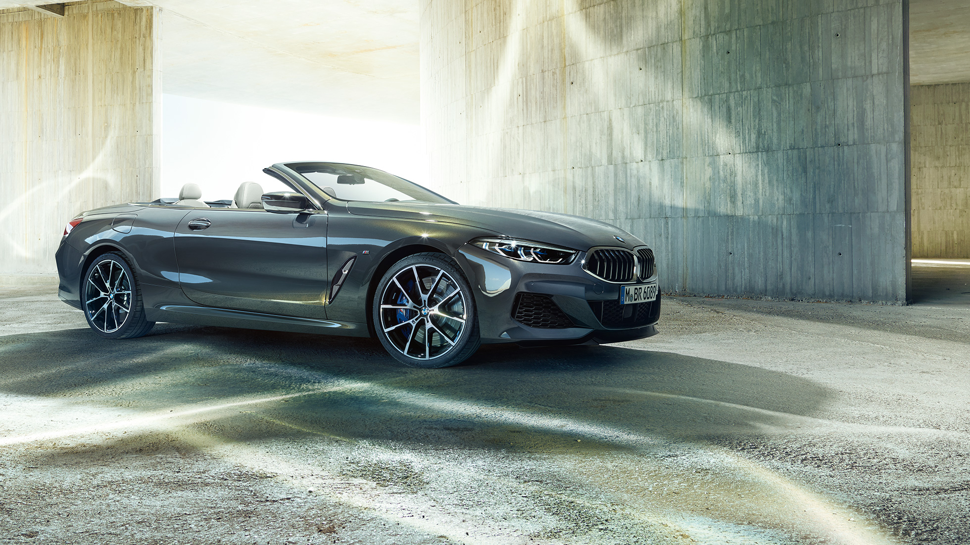 BMW M850i xDrive Cabrio, tříčtvrteční pohled z boku. BMW M850i xDrive Cabrio v šedé metalíze Dravit Grey se staženou střechou, tříčtvrteční pohled z boku, stojící před betonovou budovou.