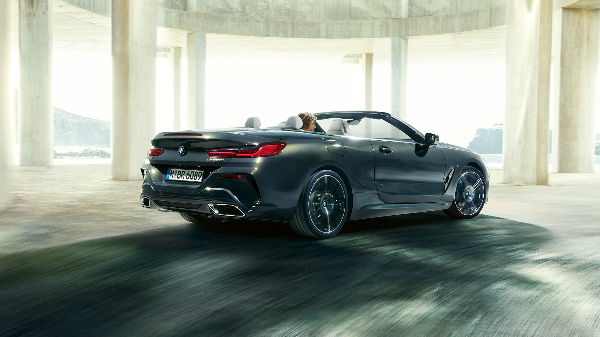 BMW M850i xDrive Cabrio, tříčtvrteční pohled zezadu. BMW M850i xDrive Cabrio v šedé metalíze Dravit Grey se staženou střechou, tříčtvrteční pohled zezadu na vůz projíždějící betonovou budovou.