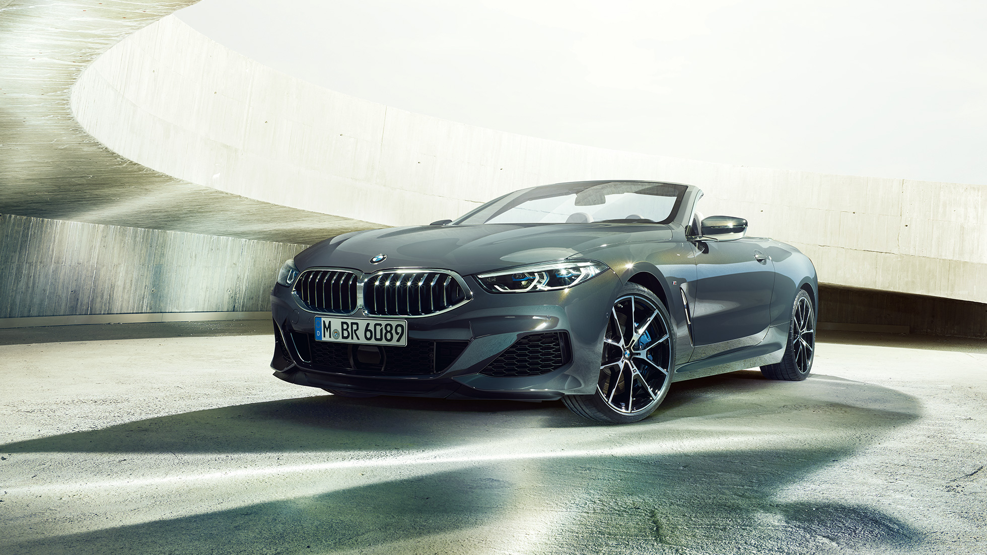 BMW M850i xDrive Cabrio, tříčtvrteční pohled zepředu. BMW M850i xDrive Cabrio v šedé metalíze Dravit Grey se staženou střechou, tříčtvrteční pohled zepředu na vůz stojící v betonové budově.