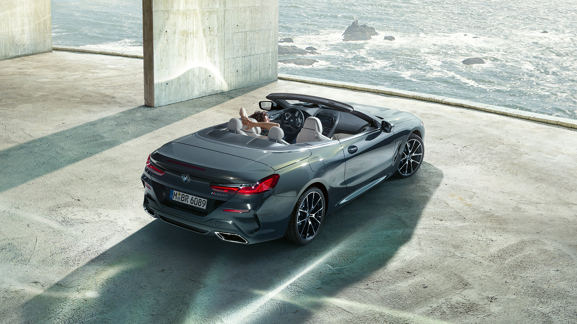 BMW M850i xDrive Cabrio, pohled z ptačí perspektivy. BMW M850i xDrive Cabrio v šedé metalíze Dravit Grey se staženou střechou, pohled z ptačí perspektivy na vůz zaparkovaný v betonové budově před oceánem.