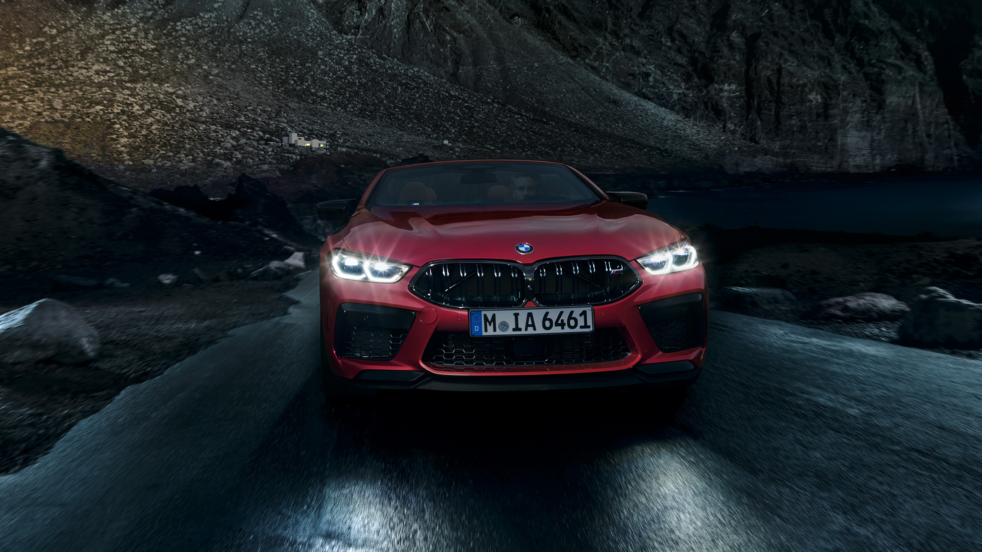 BMW M8 Competition Cabrio, pohled zepředu. BMW M8 Competition Cabrio v červené metalíze Motegi Red se zářícími předními světlomety, jízdní pohled v noci zepředu.