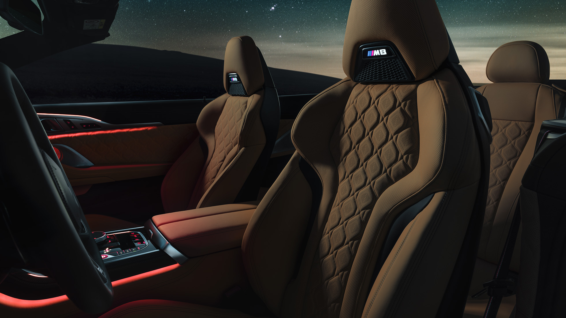 M sportovní sedadla čalouněná kůží Merino. BMW M8 Competition Cabrio se staženou střechou pod hvězdnou oblohou, interiér s M sportovními sedadly čalouněnými kůží Merino a s červeným prostorovým osvětlením.