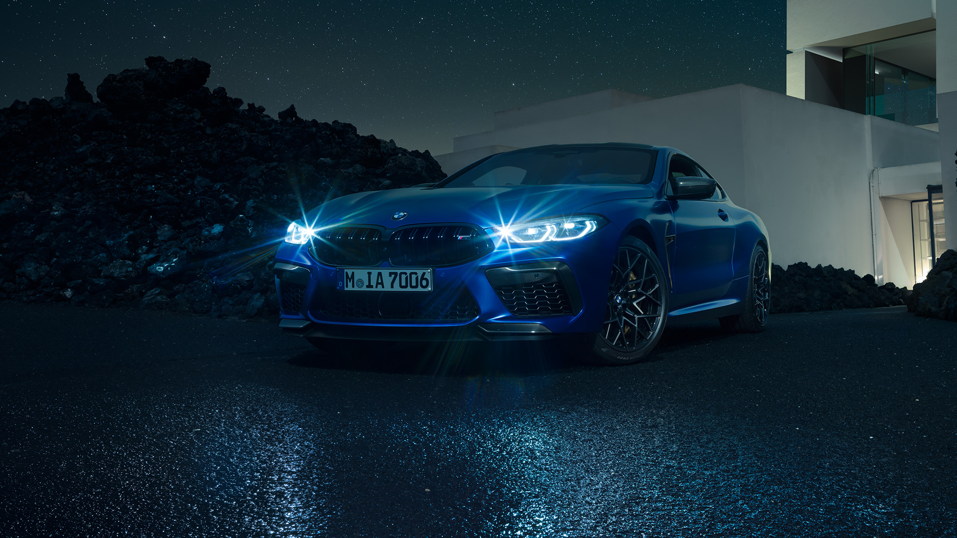 BMW M8 Competition Coupé, tříčtvrteční pohled zpředu. BMW M8 Competition Coupé v metalické barvě Marina Bay Blue s rozsvícenými LED světlomety, z tříčtvrtečního pohledu před vilou v noci.