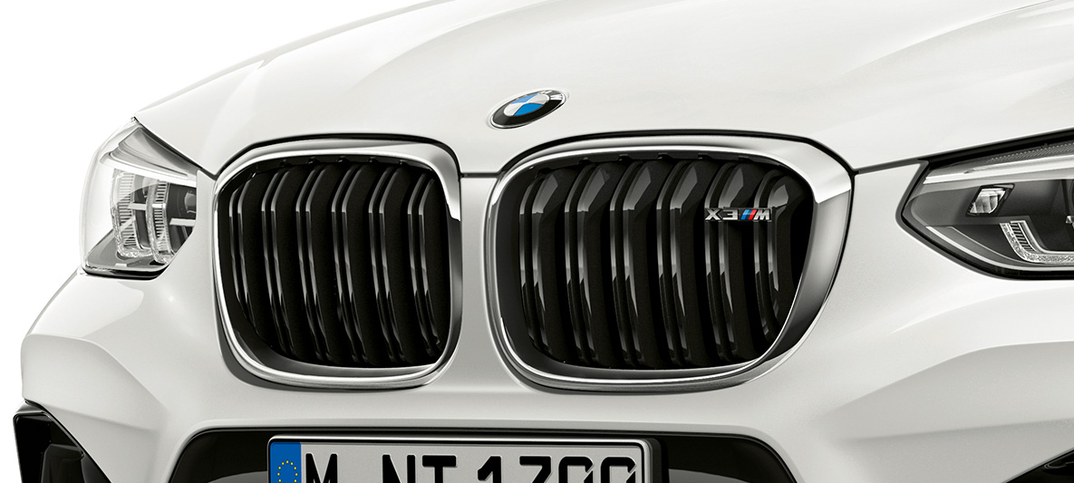 BMW X3 M, M maska chladiče ve tvaru ledvinek. BMW X3 M v bílé barvě Alpine White, exteriér, přední část, M maska chladiče ve tvaru ledvinek s chromovými detaily a označením modelu.