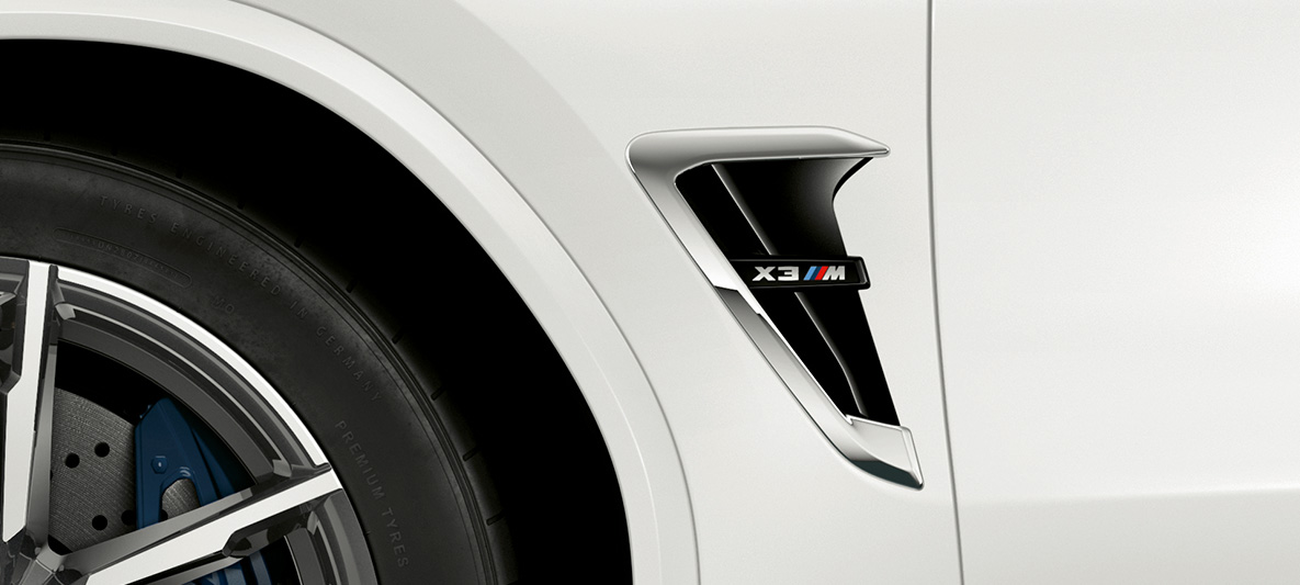 BMW X3 M, M žábry. BMW X3 M v bílé barvě Alpine White, exteriér, přední část, M žábry s chromovými detaily a označením modelu.