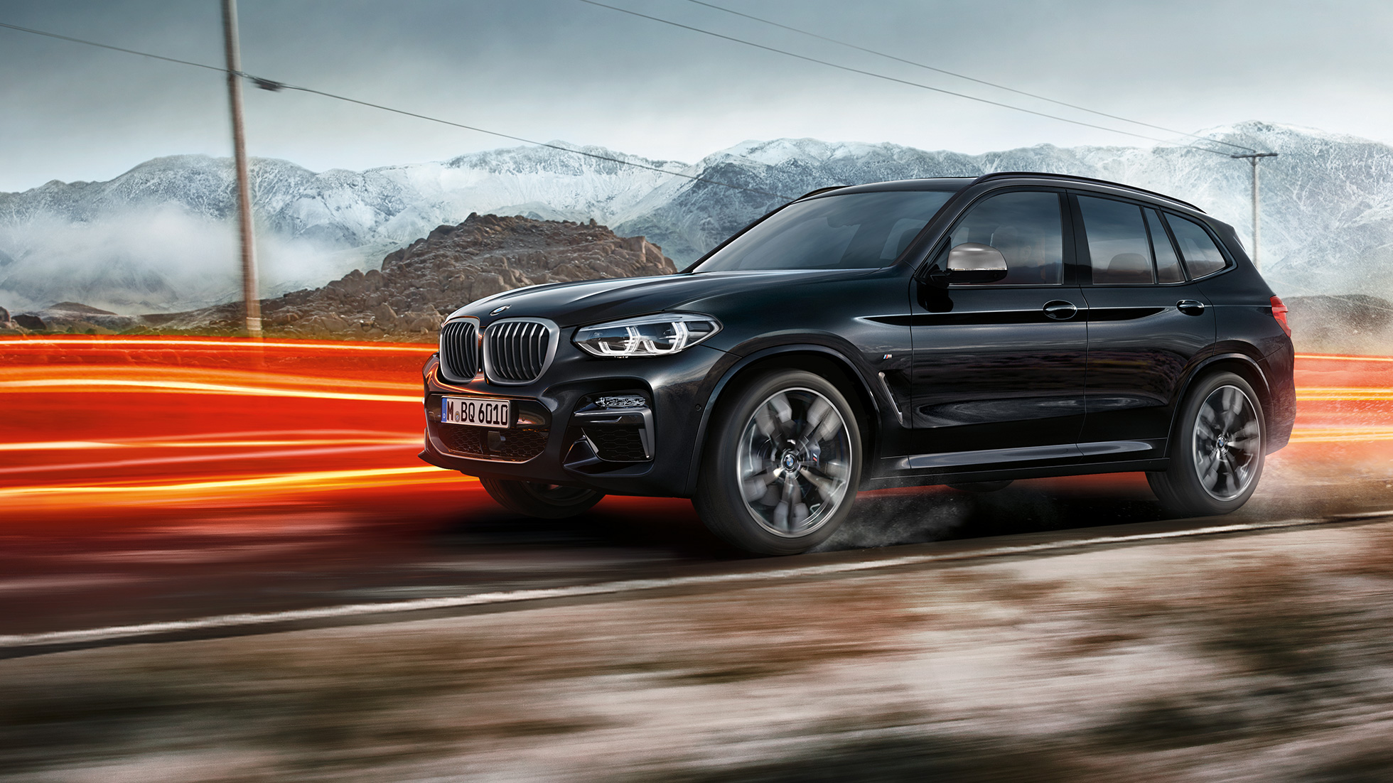 BMW X3 M40i a BMW X3 M40d, pohled zboku. BMW X3 M40i a BMW X3 M40d v černé metalické barvě Black Sapphire, jízda, exteriér, pohled zboku, v pozadí horská krajina se světelnými proužky.
