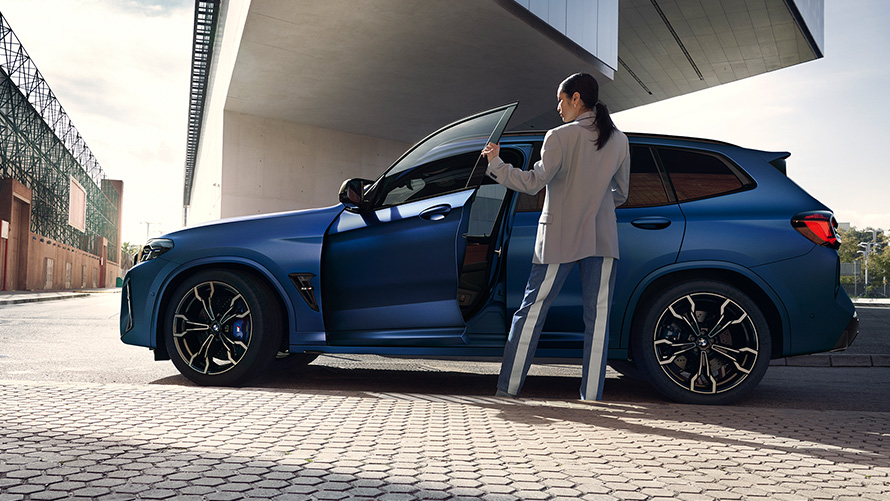 BMW X3 M Competition F97 LCI Facelift 2021 z bočního pohledu s otevřenými dveřmi a modelkou BMW X3 M Competition F97 LCI Facelift 2021 v metalické barvě Frozen Marina Bay Blue (modrá) z bočního pohledu s modelkou