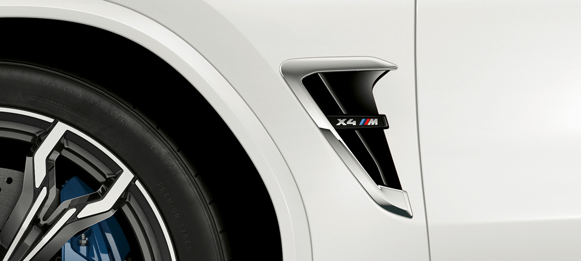 BMW X4 M, M žábry. BMW X4 M v bílé barvě Alpine White, exteriér, přední část, M žábry s chromovými detaily a označením modelu.