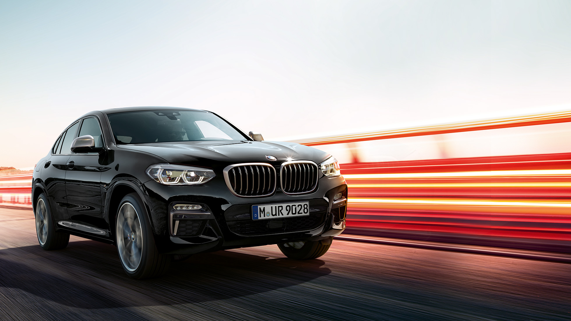BMW X4 M40i, tříčtvrteční pohled zpředu. BMW X4 M40i v černé metalické barvě Black Sapphire, jízda, exteriér, tříčtvrteční pohled zpředu se světelnými proužky.