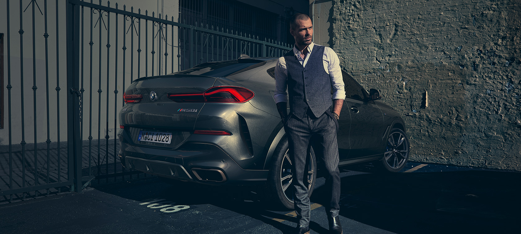 BMW X6 M50i z tříčtvrtečního pohledu zezadu. Muž stojící vedle BMW X6 M50i z tříčtvrtečního pohledu zezadu.