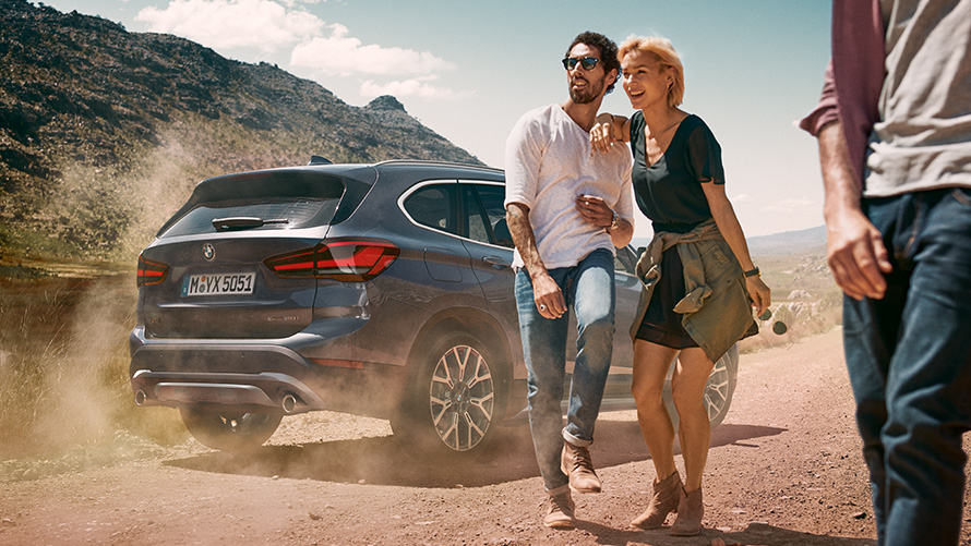 BMW X1 F48 LCI šedá Storm Bay tříčtvrteční záběr zezadu BMW X1 F48 LCI šedá Storm Bay tříčtvrteční statický záběr zezadu před krajinou s lidmi vpředu