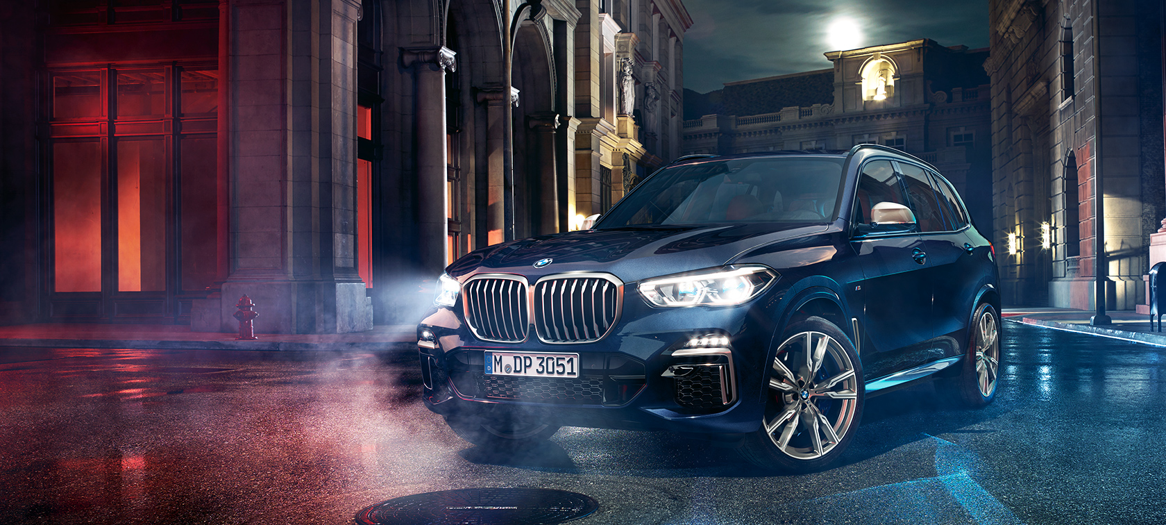 BMW X5 M50d na silnici Noční záběr BMW X5 M50d stojícího na ulici s městem v pozadí