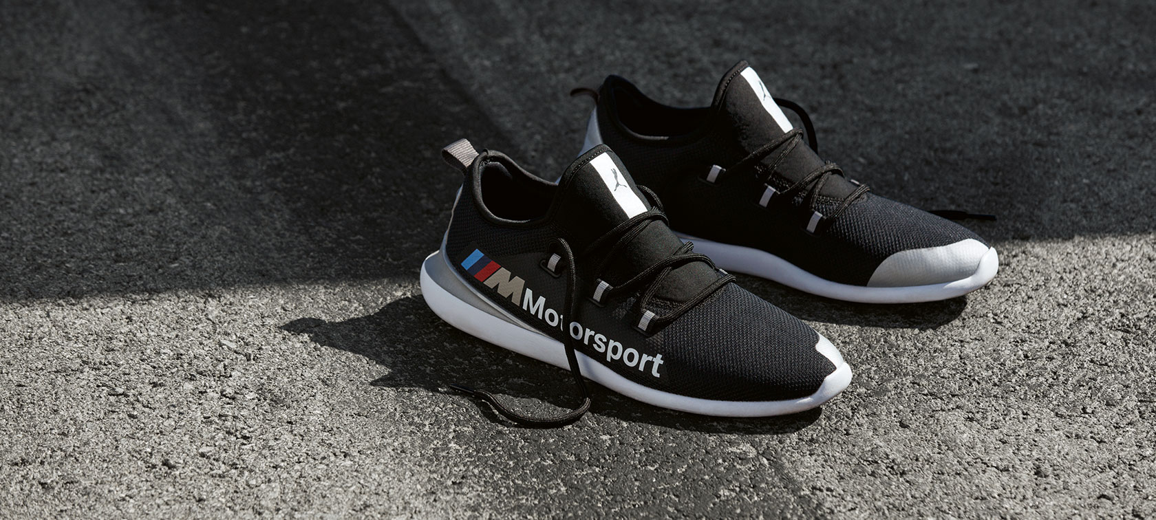 Boty BMW M Motorsport PUMA EVO CAT. Na asfaltovém povrchu leží pár bot BMW M Motorsport PUMA EVO CAT z kolekce BMW M Motorsport.